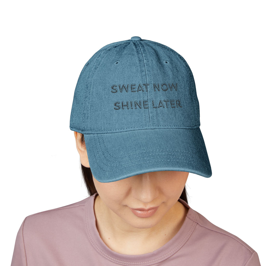 Blue denim cap with 'SWEAT NOW SHINE LATER' text worn by a person on a white background