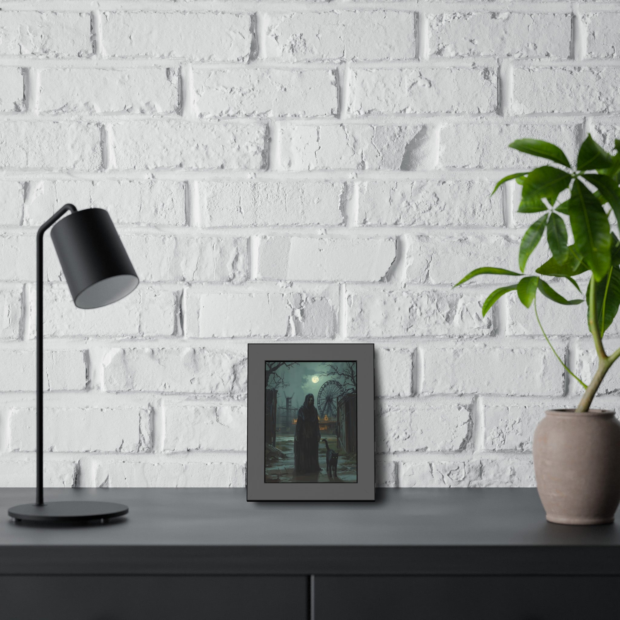 Framed artwork on a table with a lamp and plant against a white brick wall
