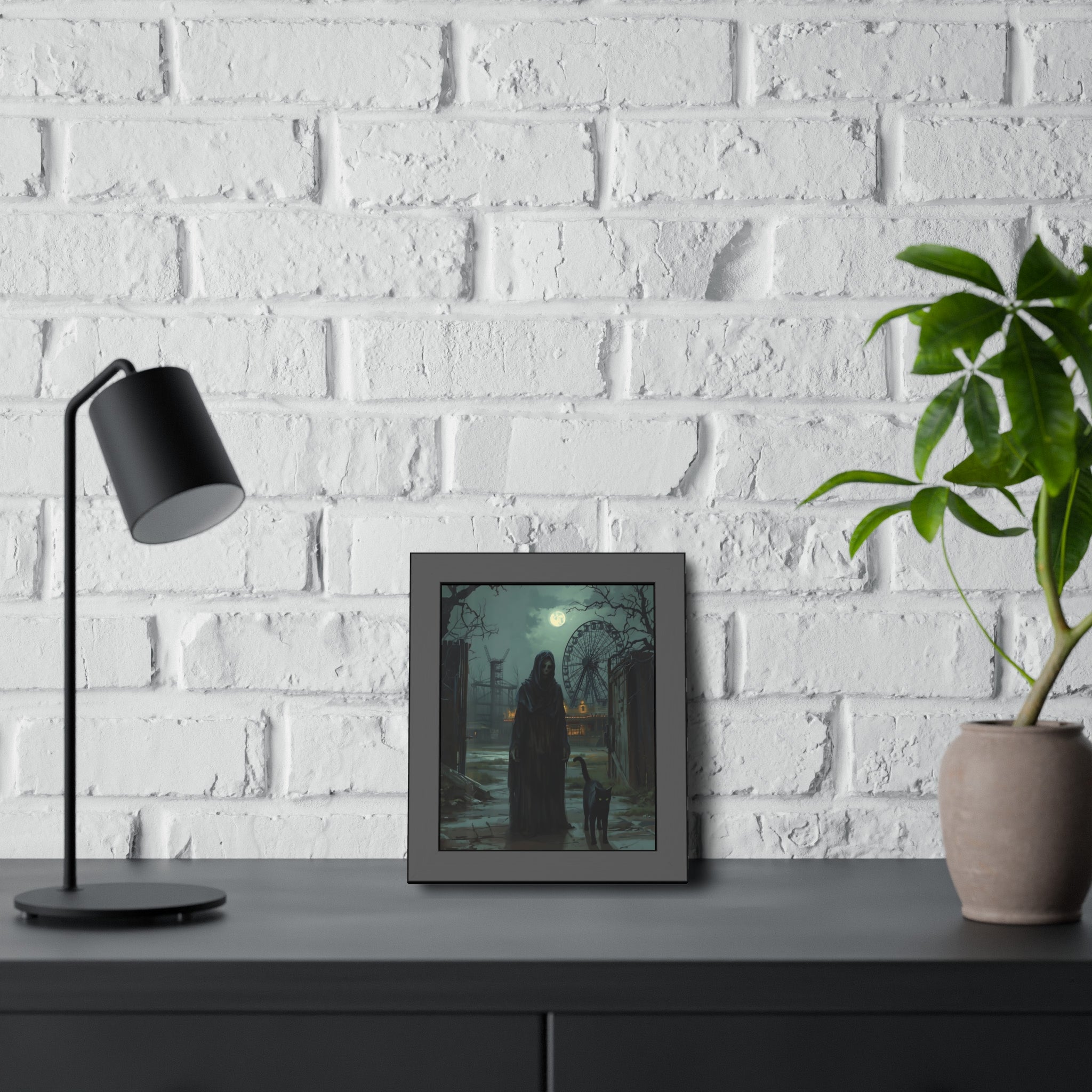 Framed artwork on a desk with a lamp and plant against a white brick wall.